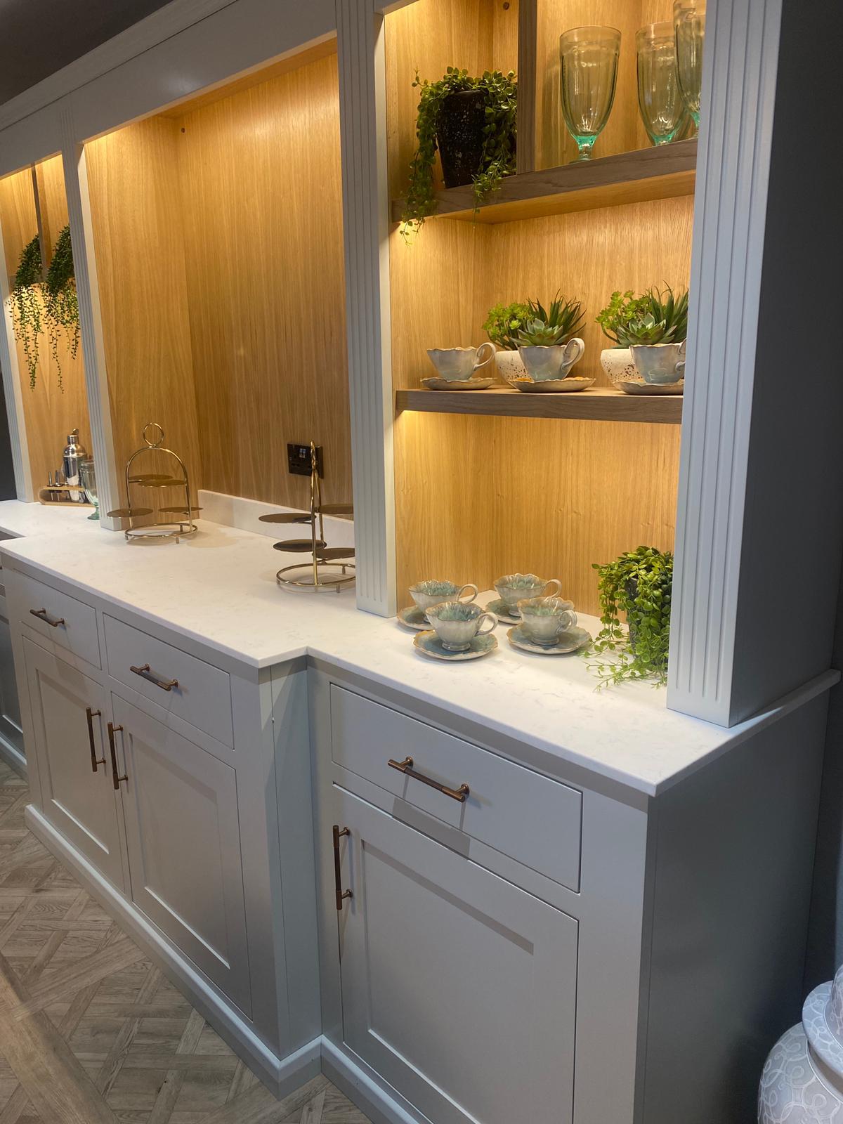 Wide view of the café display cabinetry at Meadowbrook Manor showing the full extent of the bespoke illuminated oak and grey unit with teacups, plants and glassware
