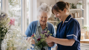 Older woman and professional carer arranging flowers together in a bright kitchen or sunroom